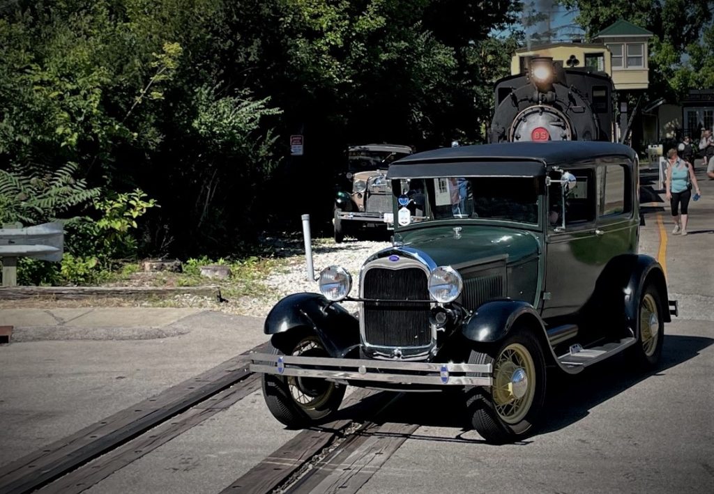 an antique ford model a tudor coupe crossing railroad traccks in front of a steam locomotive